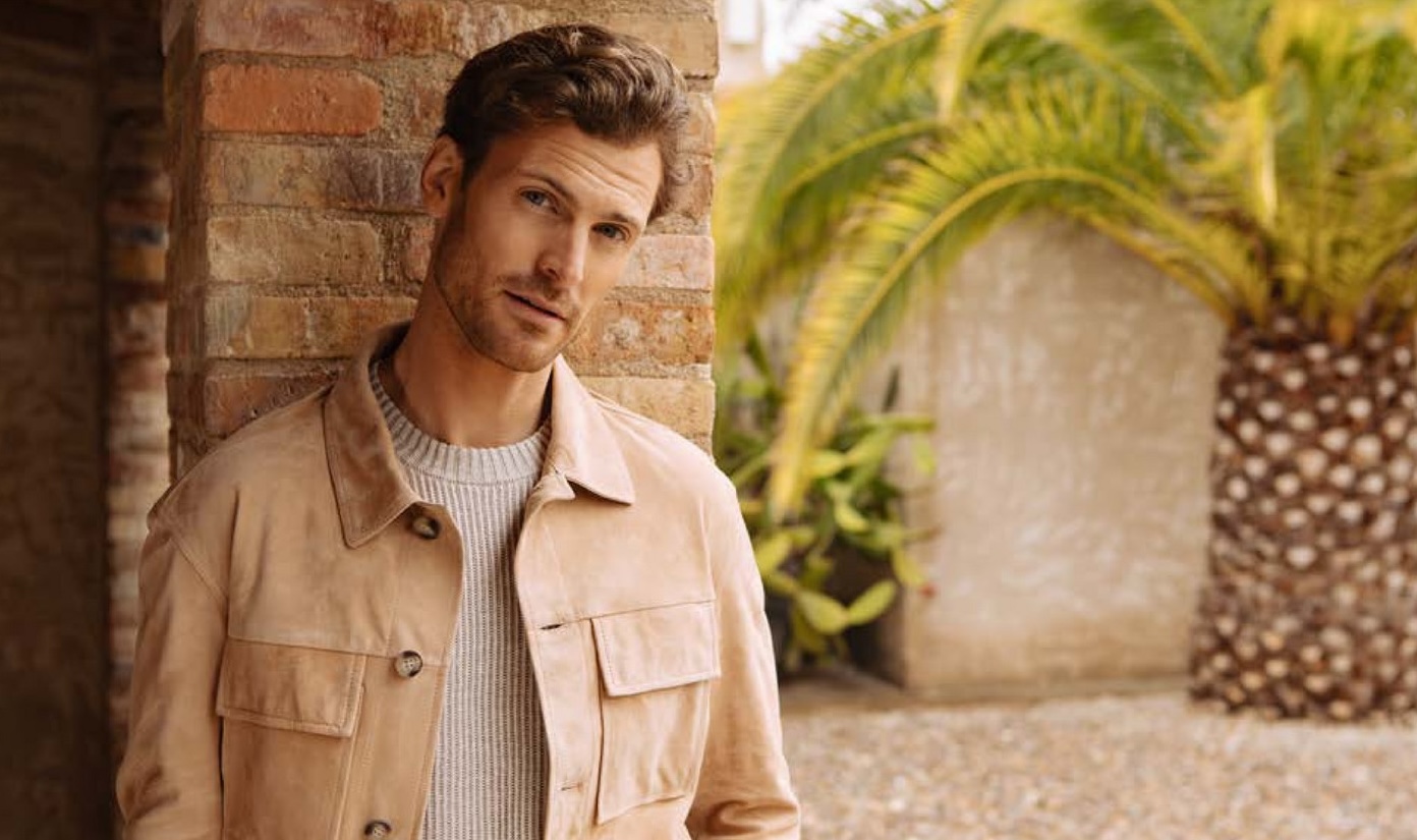 A model wearing a light brown jacket and a beige sweater of Oger, leaning against a brick wall with palm trees in the background.